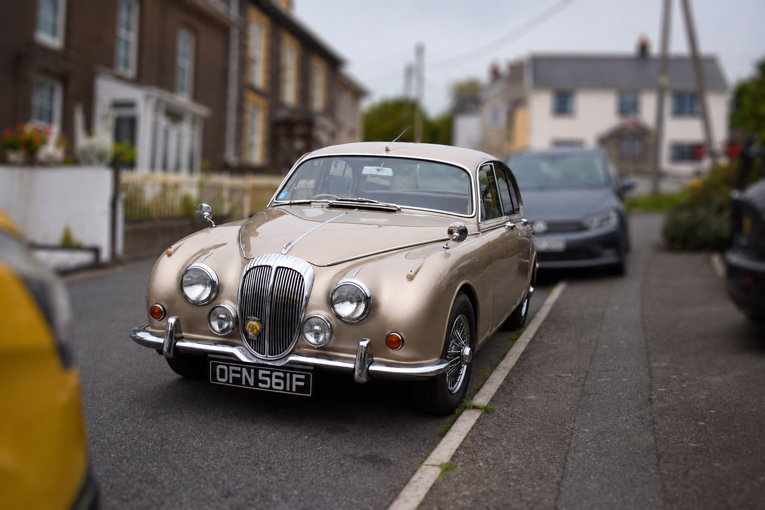 Auto-generated description: A classic beige car is parked on a residential street with houses and other vehicles in the background.