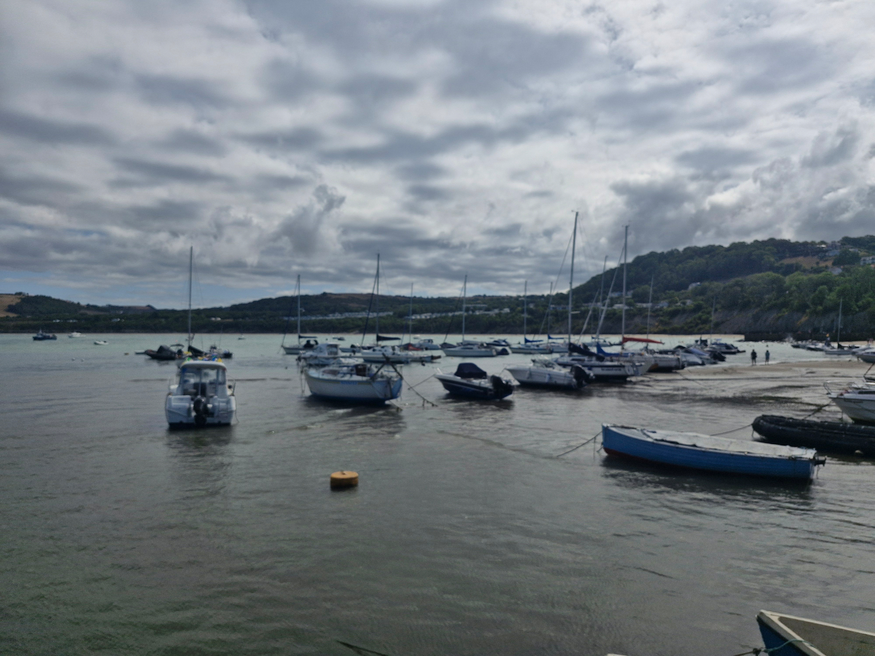 Several boats are anchored in a calm bay with cloudy skies overhead and hills in the background.
